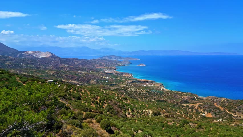 Aerial view of the rugged coastline of Crete, Greece, featuring rolling hills covered in Mediterranean vegetation meeting the deep blue sea under a clear sky in Greece.