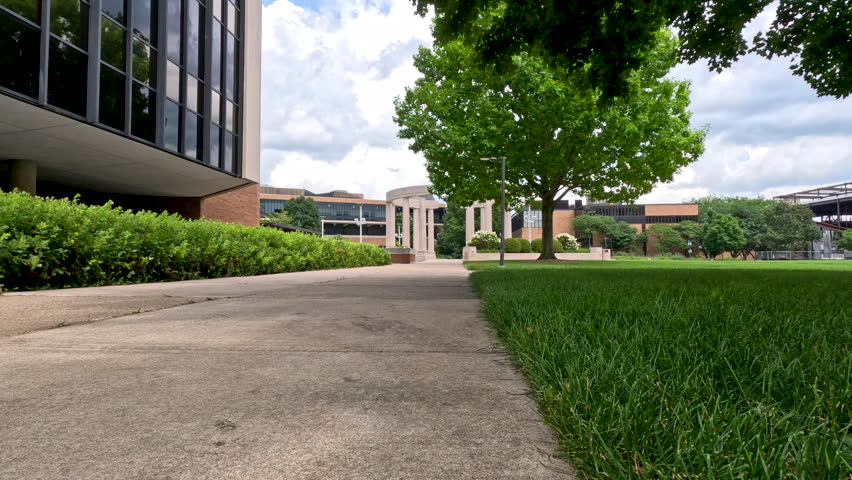 Sidewalk View Across Campus Quad with Grass Blowing in Wind. Ground-level shot looking across a grassy quad toward university buildings and a central stone colonnade with lawn grass moving gently in the breeze. Captured at University of Illinois Springfield.