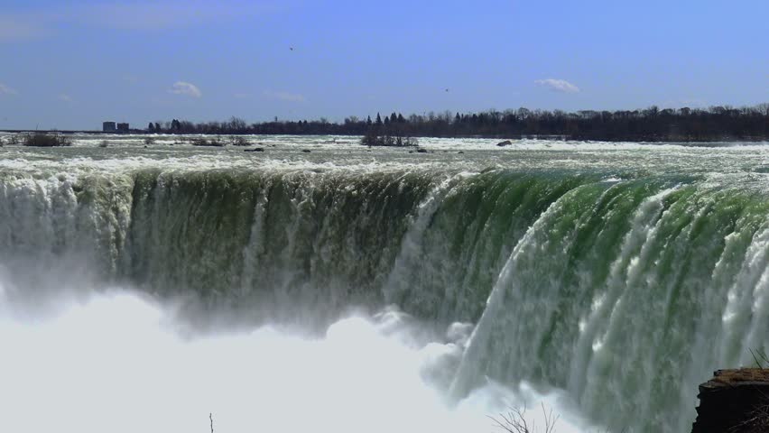 Niagara Falls on the border between Canada and the United States. The view shows the powerful flow of the Niagara River cascading over the cliff. The greenish coloration of the water is caused by a high concentration of suspended rock particles. This waterfall complex consists of three waterfalls, including the Canadian Falls, also known as Horseshoe Falls.