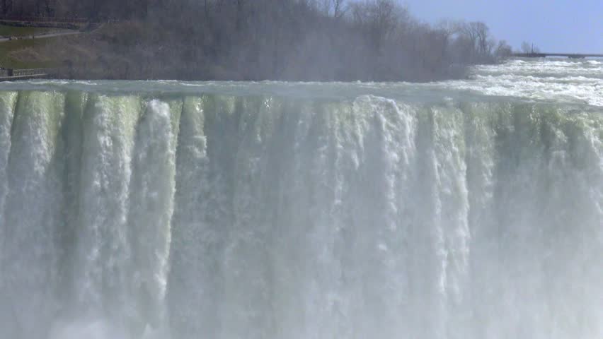 Niagara Falls viewed from the Canadian side, with the Rainbow Bridge connecting Canada and the United States visible in the background. The scene was captured in spring, with deciduous trees along the riverbank still lacking foliage.