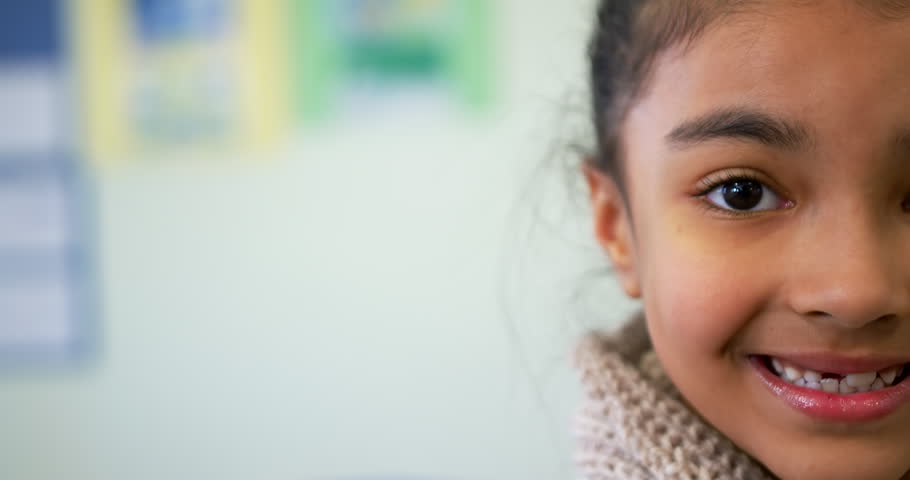 African American child with knit scarf turning to camera, showing gap smile at school, copy space. Girl, classroom, portrait, candid, cheerful, bokeh, softfocus