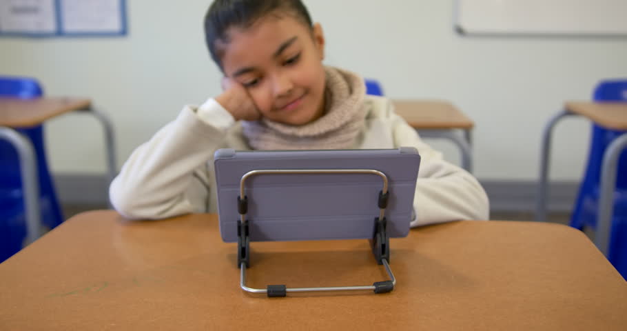 African American female child leaning on right hand in class watching tablet content causing smile. Student, screen, technology, classroom, desks, learning, education