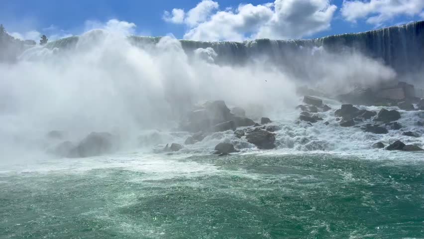 Beautiful view on the Niagara Falls and the Niagara River. Canada. Waterfall flows powerfully over rocks, creating mist and splashes, under a bright blue sky with fluffy clouds in the background.