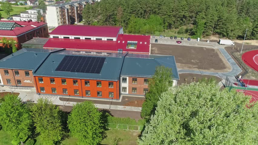 Aerial view of a modern school building with a red roof and solar panels surrounded by green trees and sports facilities in Latvia.