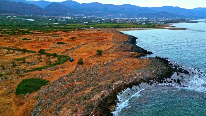 Aerial view of the rugged coastline of Crete with rocky terrain, sparse vegetation, a dirt track, and a vehicle near the sea in Greece.