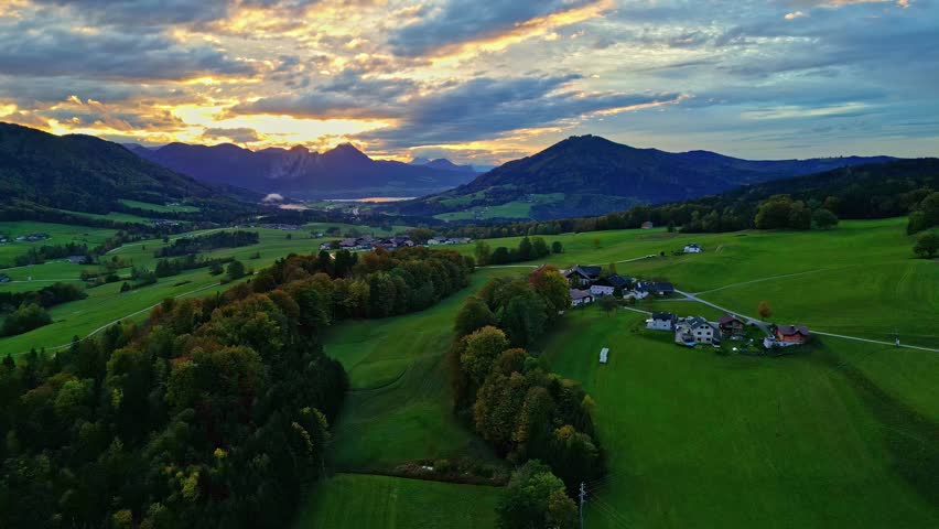 Aerial view of Attersee surrounded by lush green fields, dense forests, and rolling mountains under a dramatic sunset sky in Vocklabruck District, Upper Austria, Austria.