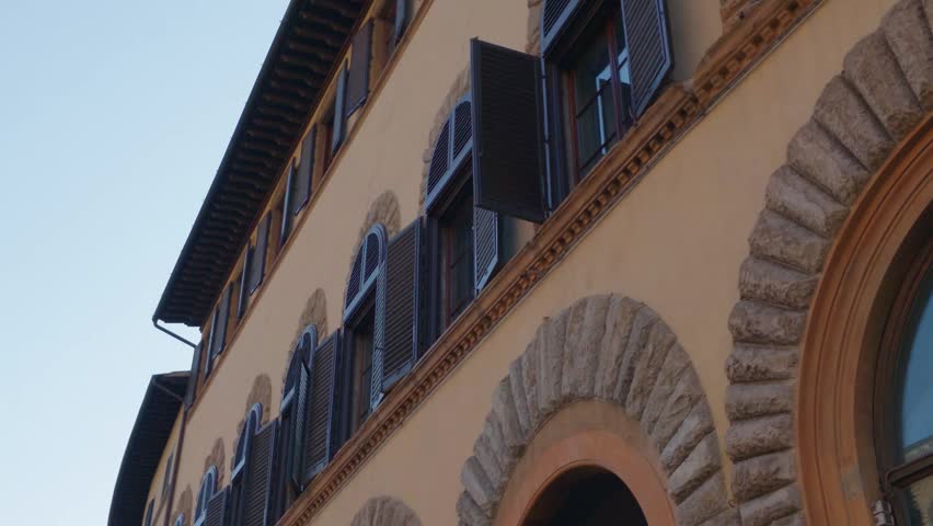 Low-angle view of a historic Renaissance building facade in Florence, Italy, featuring rusticated stone arches, traditional wooden shutters, and elegant Tuscan architecture under a clear blue sky.
