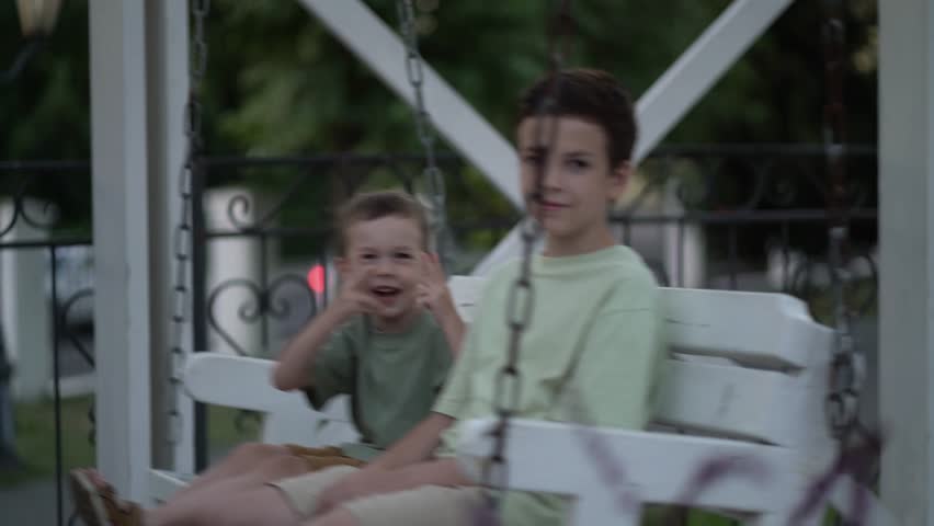 Two brothers swinging on a white porch swing in a quiet outdoor park, smiling and sharing a carefree childhood moment together.