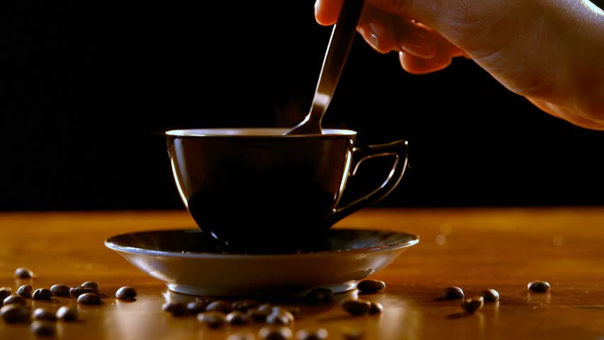 Right hand entering frame and stirring black cup with metal teaspoon, creating concentric ripples. Coffee, ceramic, wood, beans, warmth, studio, minimalist