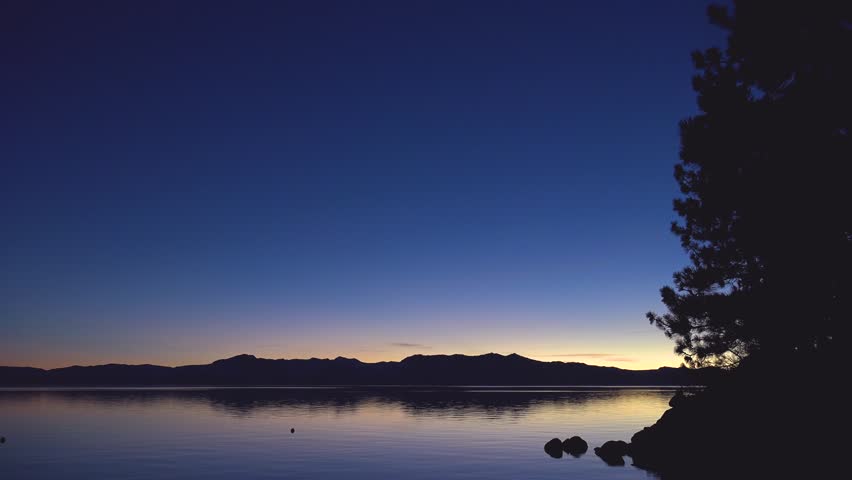 Peaceful lake scenery during dusk with silhouetted trees and hills.