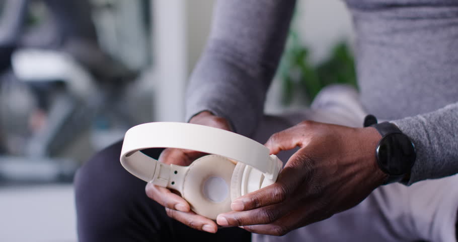 African man in gray top picking up, inspecting white headphones in gym, adjusting fit for workout. Headset, wireless, earcups, minimalist, focus, homegym, ambient