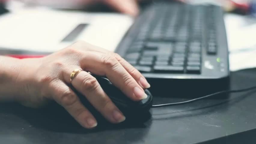 Hand holding mouse while fingers type on keyboard in busy office setting with scattered papers and technology devices. Focus on work activity and productivity in modern workspace.