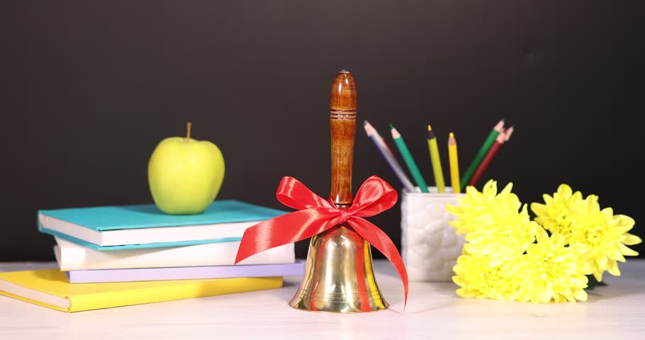 Woman putting pencils into holder, school bell, books, apple and flowers at white wooden table near chalkboard, closeup