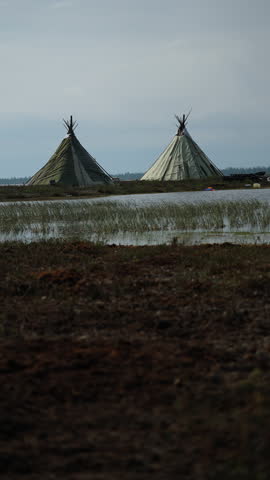 Two traditional conical tents stand beside a calm marsh in a remote tundra landscape under a cloudy sky.
