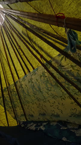 Close-up view inside a rustic tent showing bamboo poles, rope ties, and diffused light through a yellow tarp, with blue floral fabric lining and bedding. Abstract lines and cozy camping mood.