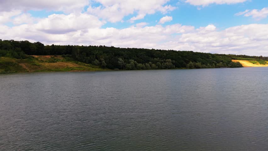 Aerial drone flying low over the water of a lake toward a wooded shoreline under a blue summer sky with white clouds