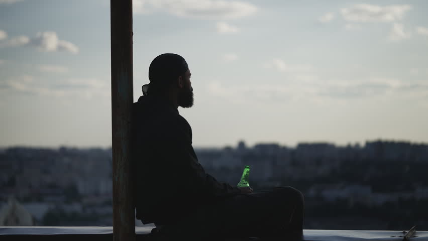 Young man in a durag sits on rooftop edge and drinks beer, lost in thoughts