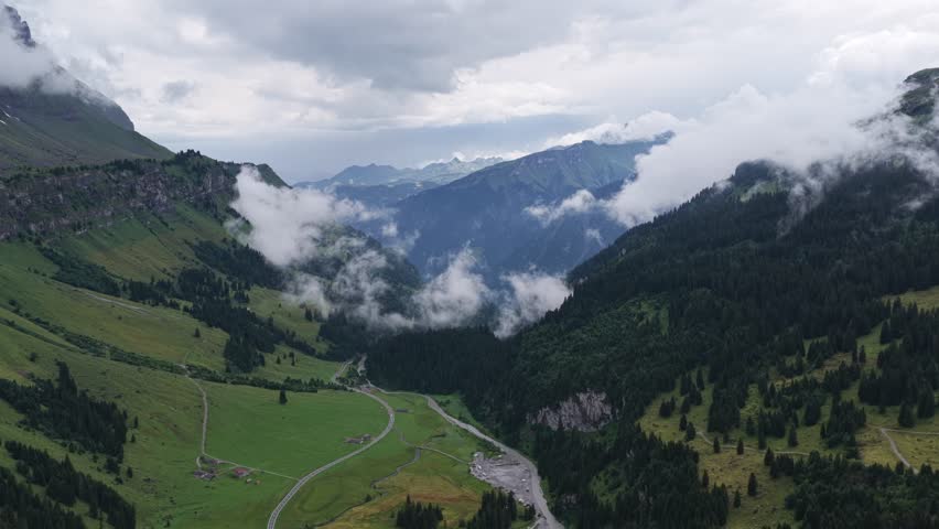 Cinematic drone footage captures a dramatic aerial view over a Swiss alpine valley surrounded by steep green slopes, dense conifer forests and distant mountain ridges. Low clouds drift gently through the landscape, creating an atmospheric and moody scene that highlights the scale and beauty of the Swiss Alps. The camera perspective looks deep into the valley, where a winding mountain road and a narrow riverbed guide the eye through the composition and add a strong sense of depth and natural structure.