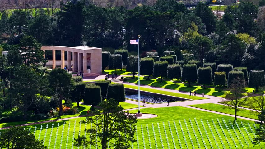 Aerial view of the Normandy American Cemetery and Memorial with rows of white crosses, manicured trees, and a reflecting pool in Colleville-sur-Mer, France.