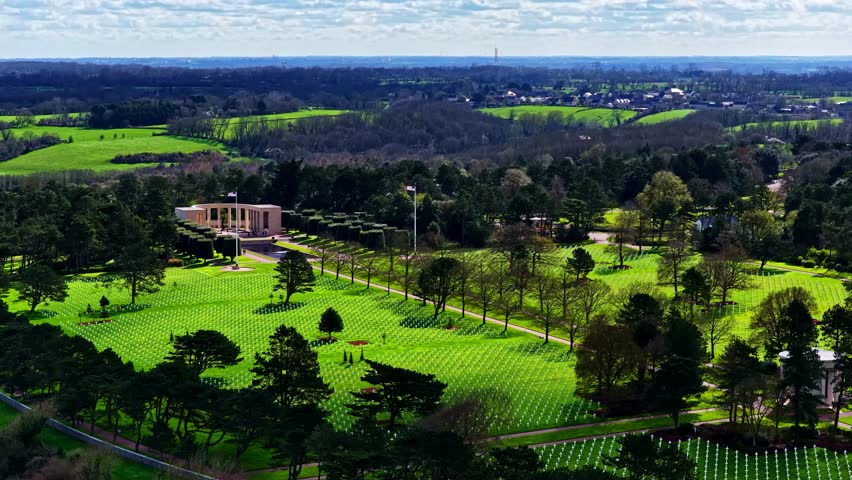 Aerial view of the Normandy American Cemetery and Memorial with rows of white crosses set in green lawns surrounded by trees in Colleville-sur-Mer, France.