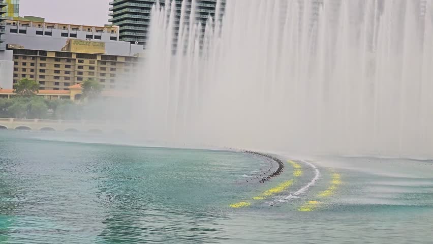 Bellagio Fountains performing on the Las Vegas Strip, with rows of water jets, misty spray, blue green lake, resort architecture, city skyline, and bright daytime scenery.