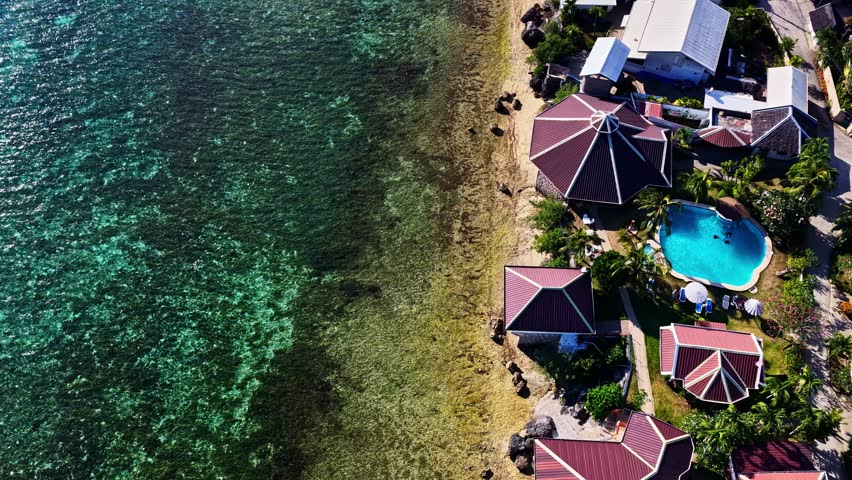 Aerial view of a tropical resort featuring a swimming pool and buildings with dark red roofs situated along the clear turquoise waters of the Philippines.