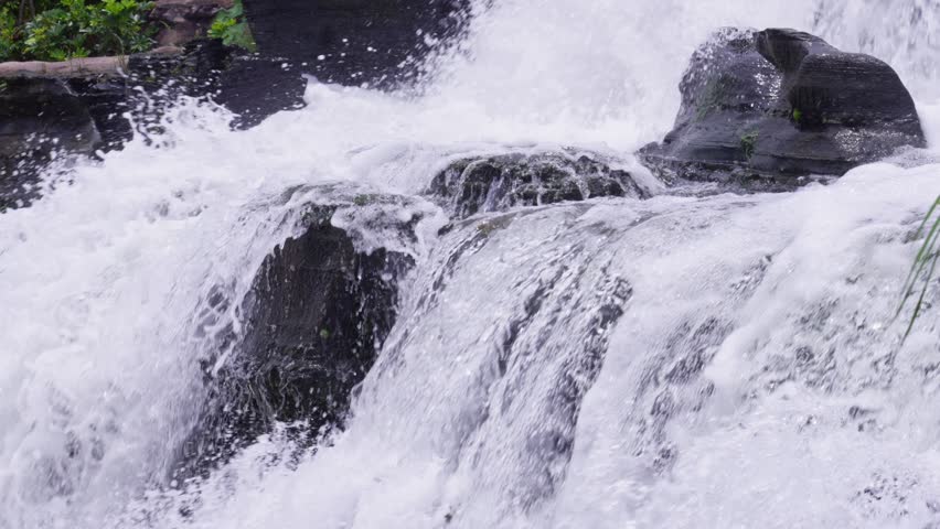 A close-up view of a garden waterfall where white water flows quickly over dark rockery rocks, splashing continuously and forming fine water splashes.