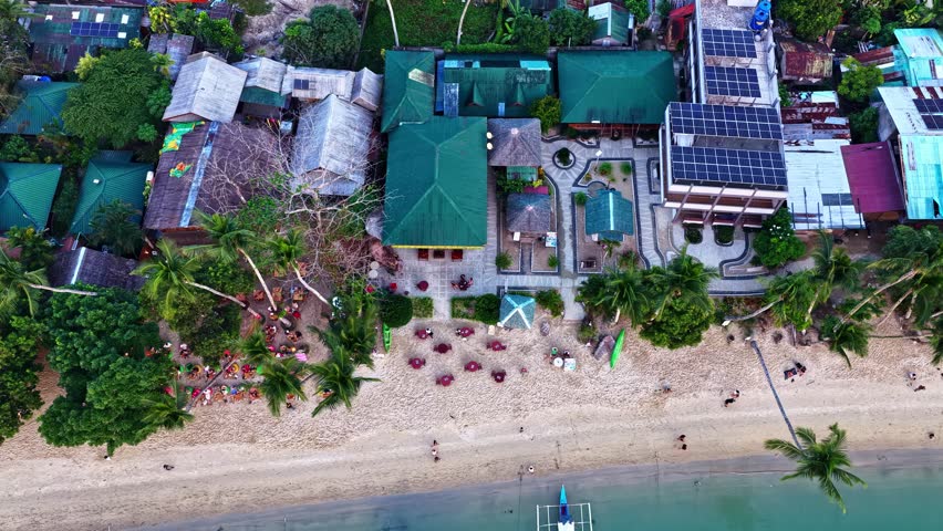 Aerial view of a tropical beach resort with green-roofed buildings, solar panels, and palm trees along the sandy coastline in the Philippines.