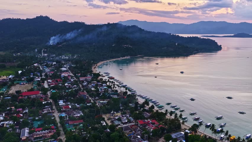 Aerial view of a coastal town with houses, boats along the beach, and hills under a sunset sky in the Philippines.