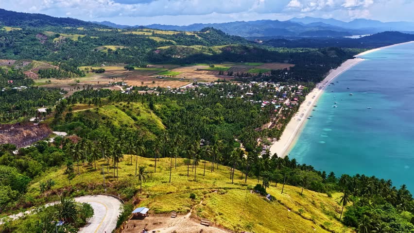 Aerial view of a coastal landscape featuring lush green hills, palm trees, a winding road, and a sandy beach meeting the turquoise sea in the Philippines.