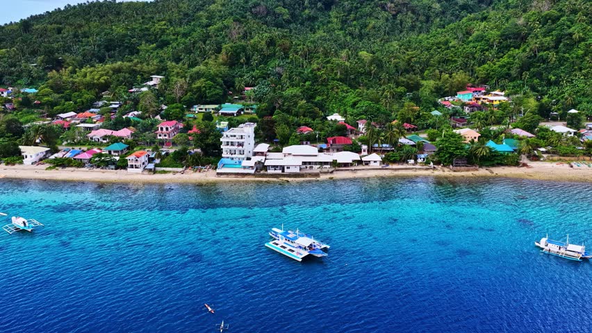 Aerial view of a coastal village with colorful houses, lush green hills, and traditional outrigger boats floating on clear blue water in the Philippines.