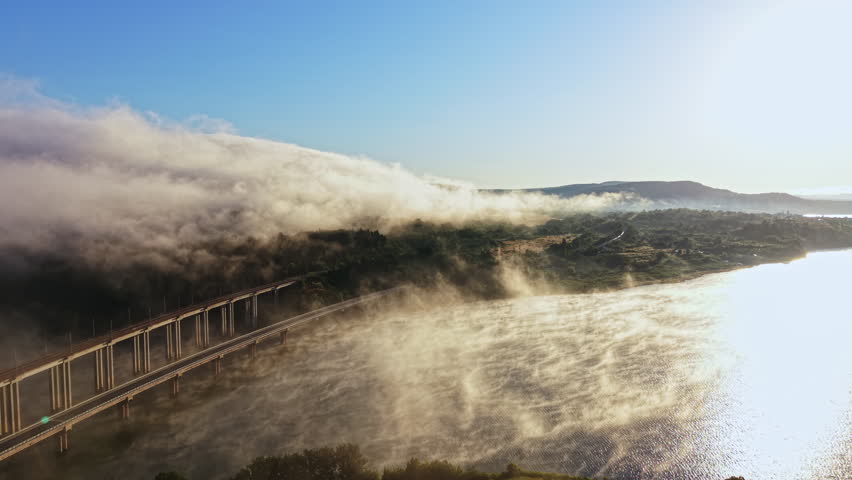Fog drifts over the river and the surrounding hills as sunlight breaks through at dawn. The bridge crosses the river, adding to the view of the natural setting.