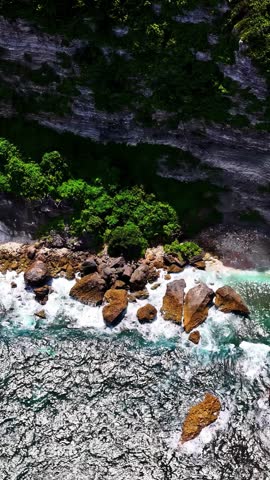 Aerial view of rugged coastal cliffs with lush green vegetation and crashing waves hitting large rocks along the shoreline in Bali, Indonesia.