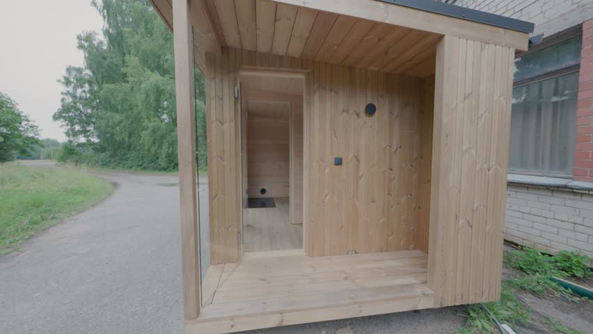 Front entrance of a compact modular timber cabin with a covered porch, glass side panel, open doorway, and natural wood cladding beside a rural road