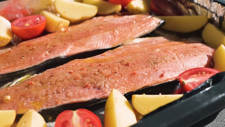 Salmon fillets are placed on a baking tray surrounded by sliced tomatoes and potatoes. The scene takes place in a kitchen, showing a food preparation process.