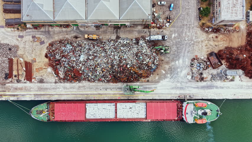 A cargo ship is docked alongside a scrapyard. The ship is taking on materials from the nearby pile of scrap metal. Trucks and machinery are present at the site.