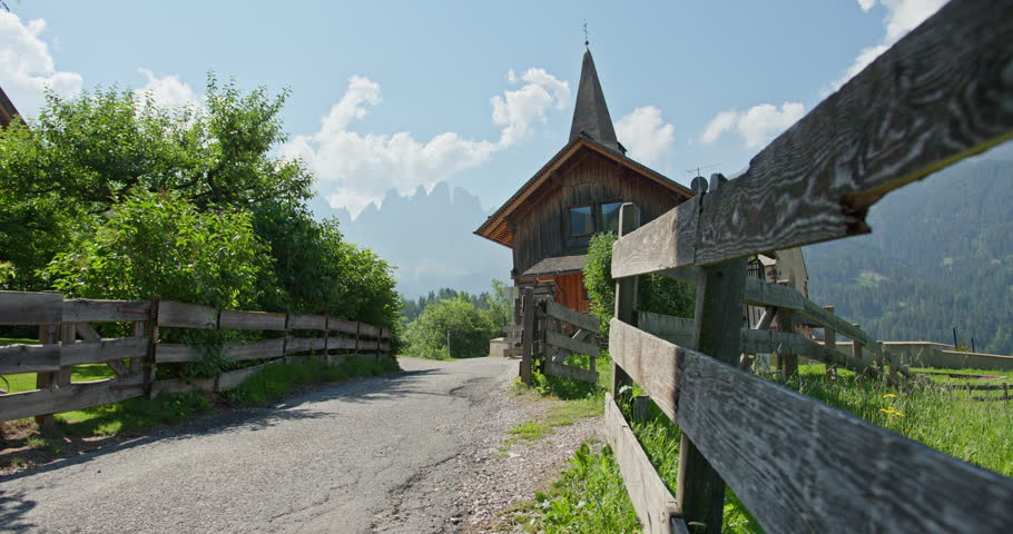 Sunny summer view of a rural lane with wooden fences, a traditional alpine house and a church steeple in Santa Maddalena, Val di Funes, with the Odle mountains in the background, South Tyrol, Italy.