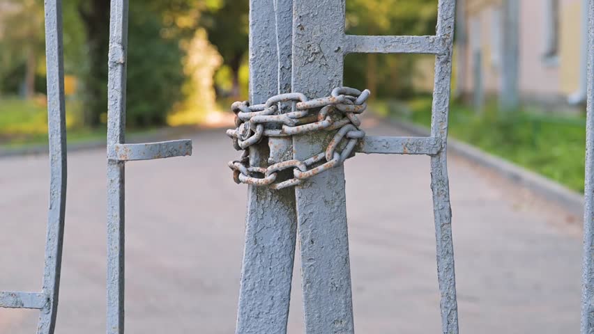 A metal gate is secured with chains, showing a close-up of the lock mechanism. The background features a blurred pathway and greenery, suggesting an outdoor area that is currently inaccessible.