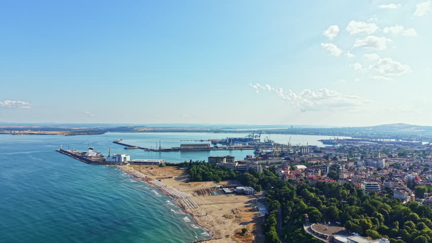 A coastal city stretches along the shore with sandy beaches and green trees. Ships are docked in the harbor while buildings line the waterfront. People enjoy the sun.
