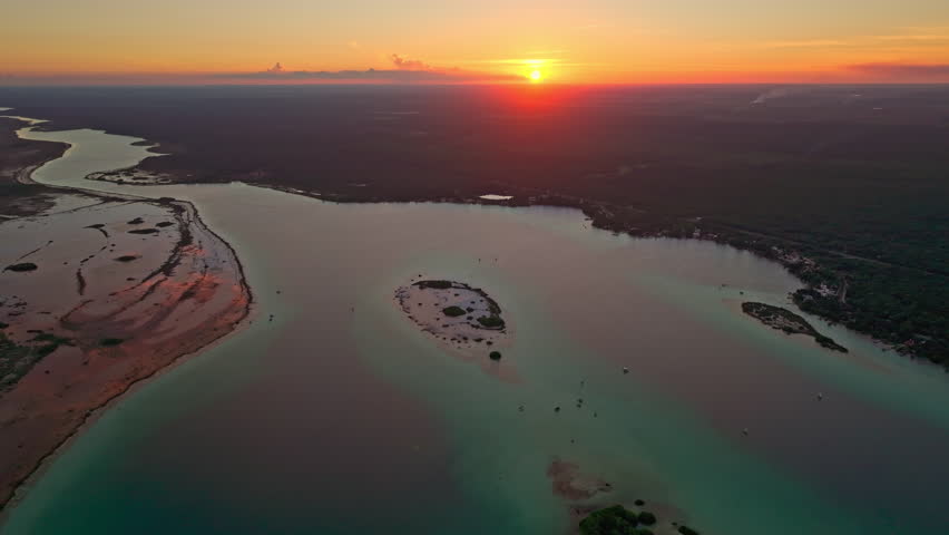 Aerial view at sunset over a winding channel of the Bacalar lagoon, Mexico, the orange sun on the horizon reflected in the calm still water.
