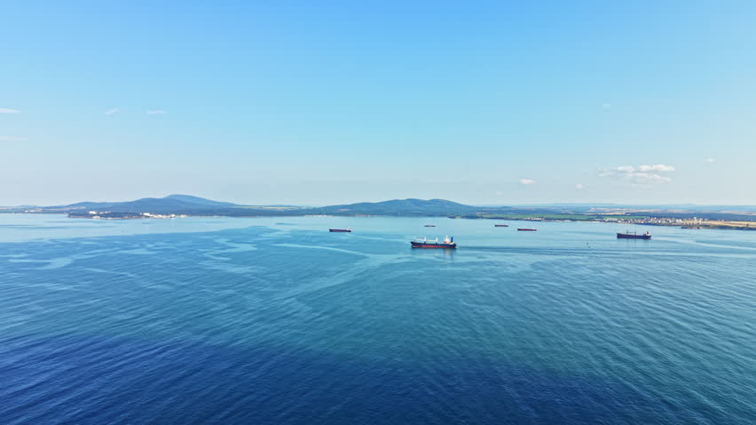 Several ships navigate the clear waters of the harbor. Mountains rise in the distance against a bright blue sky. The scene shows an active port area during daylight hours.