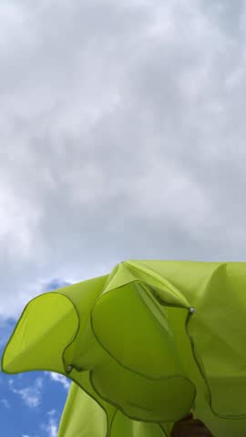 Person opening a bright green beach umbrella against a cloudy summer sky, low-angle view of setting up shade and sun protection for a relaxing holiday