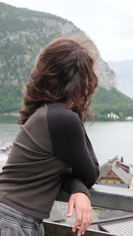 Child with windblown curly hair leaning on a railing and gazing out over a calm alpine mountain lake and village, peaceful summer travel and wanderlust moment