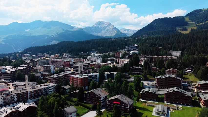 Aerial view of the Guarda Golf Hotel and Residences surrounded by alpine buildings and lush green forests in Crans-Montana, Valais, Switzerland.