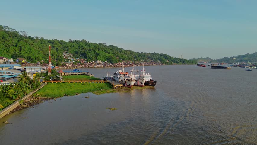 Aerial view of ships docked at a river pier with lush green hills and buildings along the bank in Samarinda, East Kalimantan, Indonesia.
