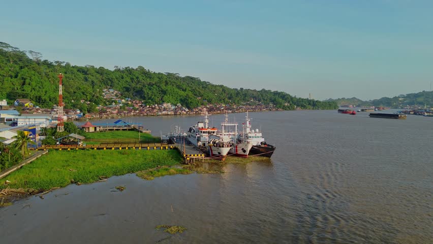 Aerial view of ships docked at a river pier with lush green hills and residential buildings in the background in Samarinda, East Kalimantan, Indonesia.