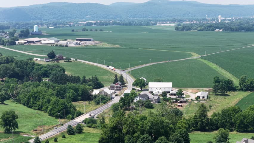 Donegal Creek farmland stretches beneath rolling ridgeline, white barn and farmhouse sit near crossroads where truck travels rural highway through Pennsylvania fields. Aerial drone wide shot