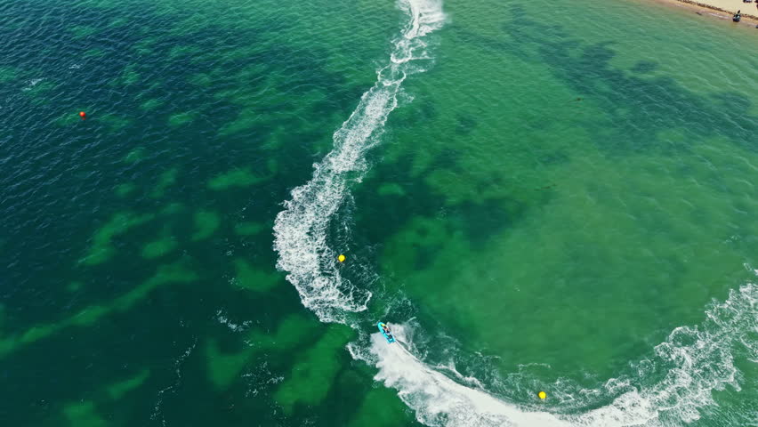 Jet ski riders race on a bright day at a coastal location. Waves form trails in the water as the participants speed by. Yellow buoys mark the race course in the distance.
