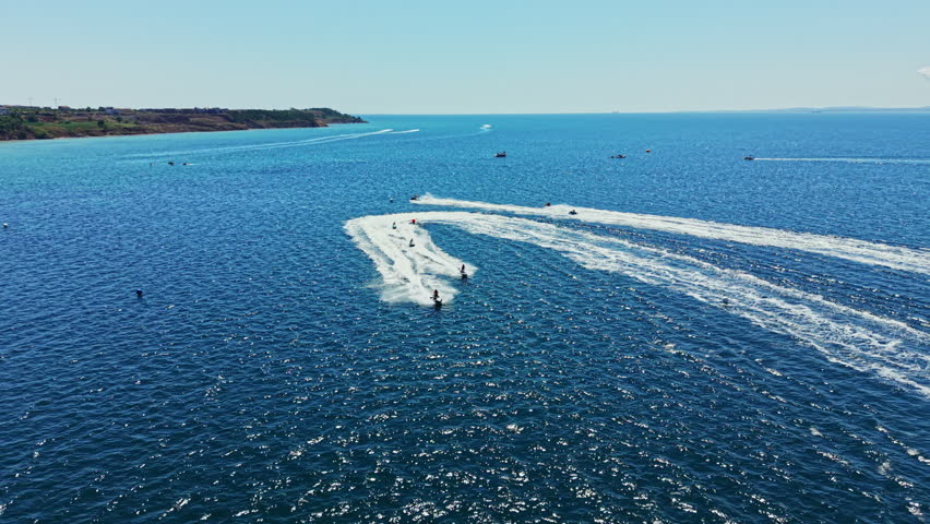 Riders on jet skis race across the water on a sunny day. The sea is blue and clear, and trails of white wake are visible behind the jet skis.