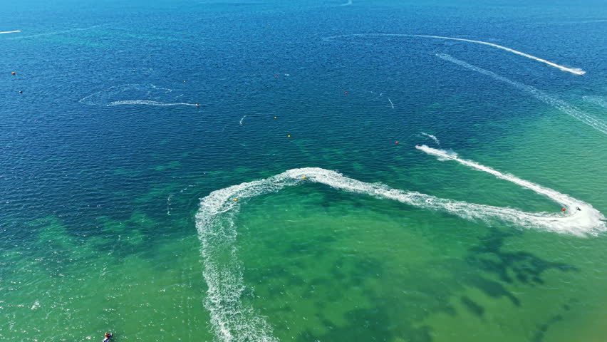 Participants race jet skis in clear blue water. The bright sun shines over the scene. Tracks from the jet skis are visible in the waves as riders enjoy the fast-paced sport.